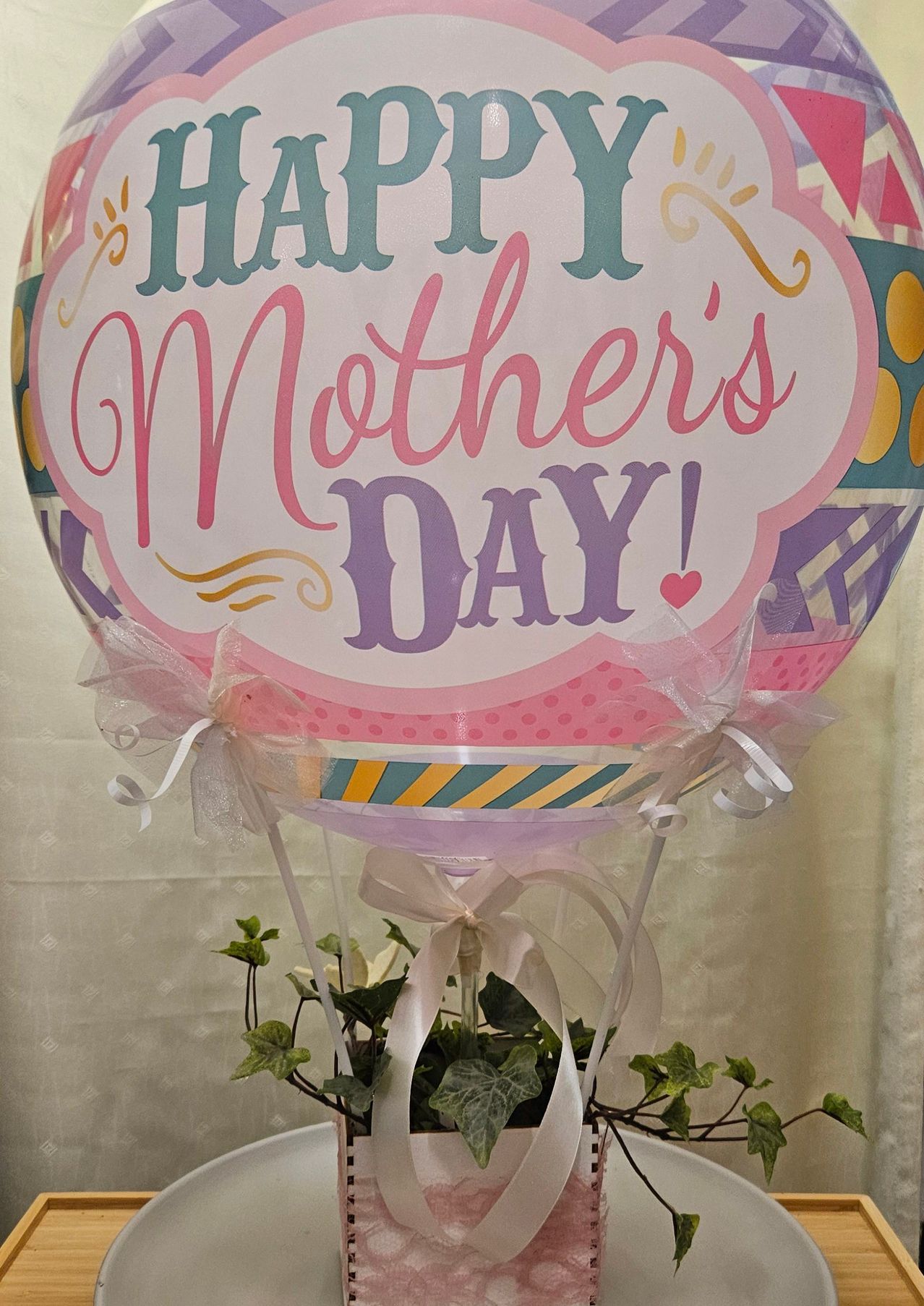 Ein farbenfroher Heißluftballon mit 'Happy Mother's Day!' in Pink und Lila beschriftet, geschmückt mit Bändern und einer Pflanze.