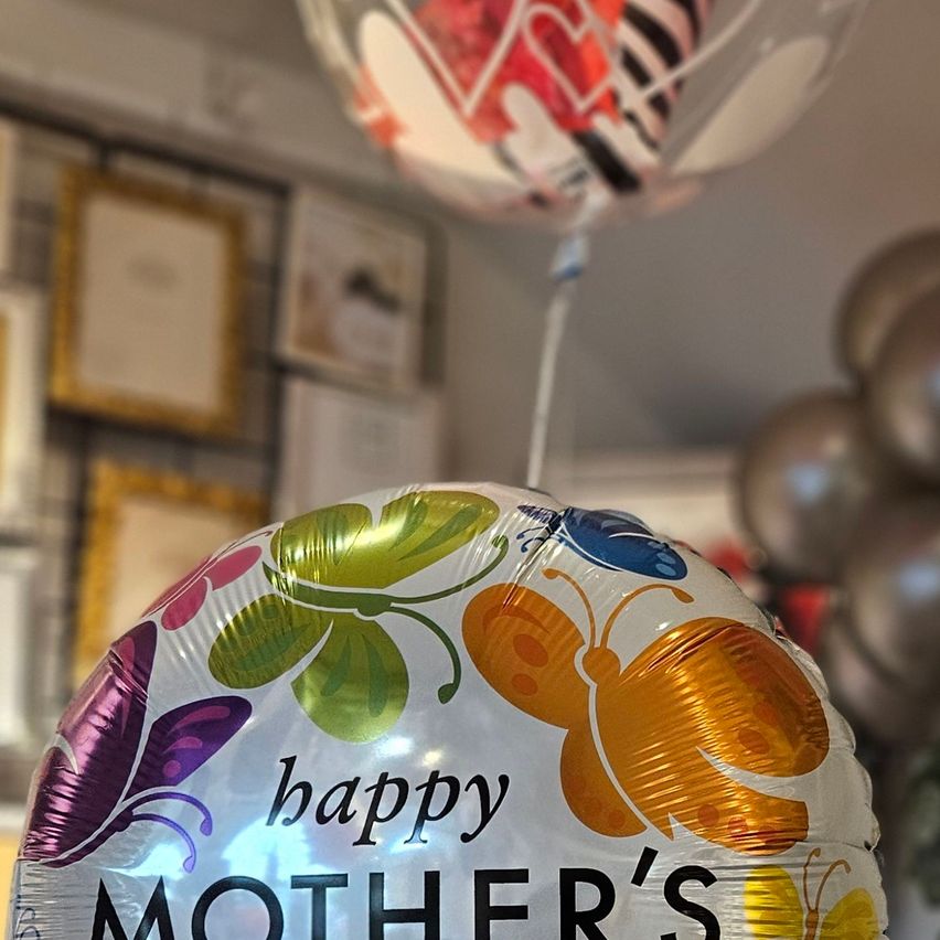 Ein bunter Muttertagsballon mit Schmetterlingen, der über einem Raum mit gerahmten Urkunden hängt.