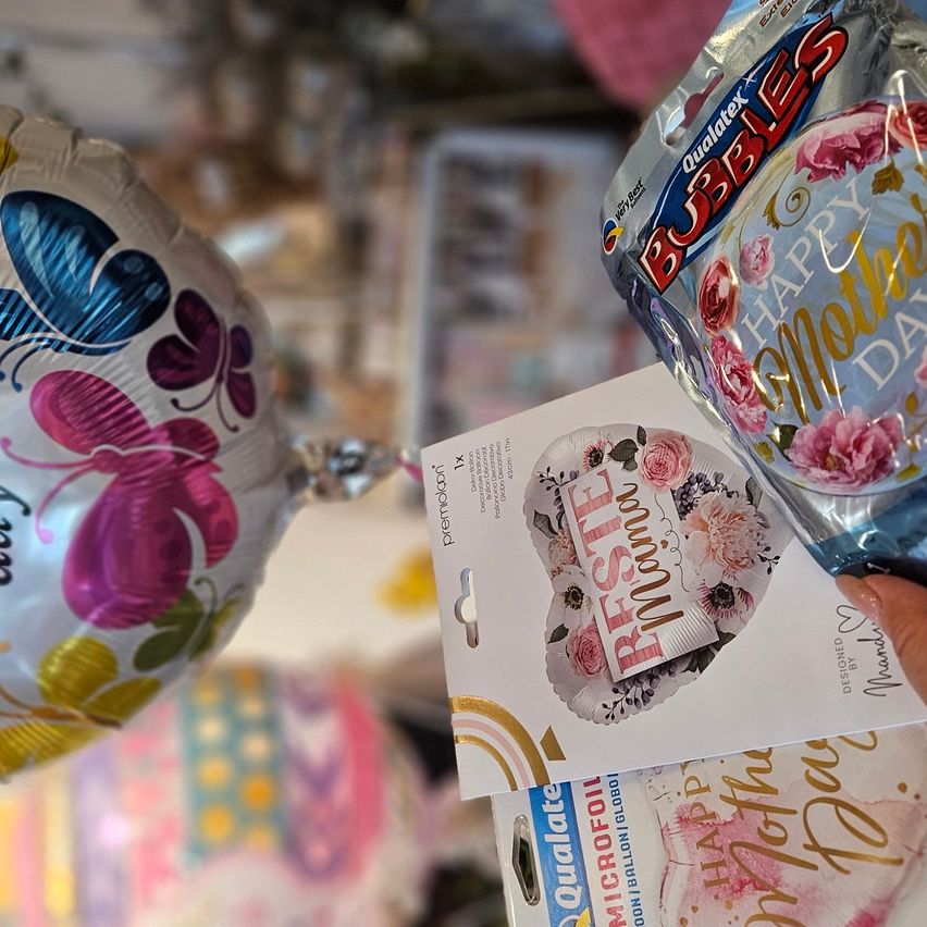 Zwei festliche Luftballons, einer mit Blumen- und einer mit Schmetterlingsdesign, werden von einer Hand gehalten. Der Blumenballon hat eine 'Happy Mother's Day'-Botschaft.