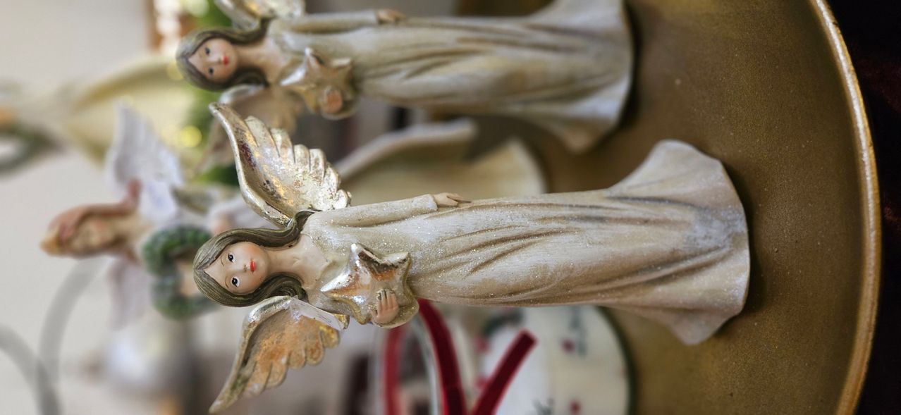 Nahaufnahme einer weißen Engelsfigur mit Flügeln, einem Stern auf der Brust und einem Anhänger am Rücken. Eine weitere Engelsfigur ist teilweise im Hintergrund zu sehen.