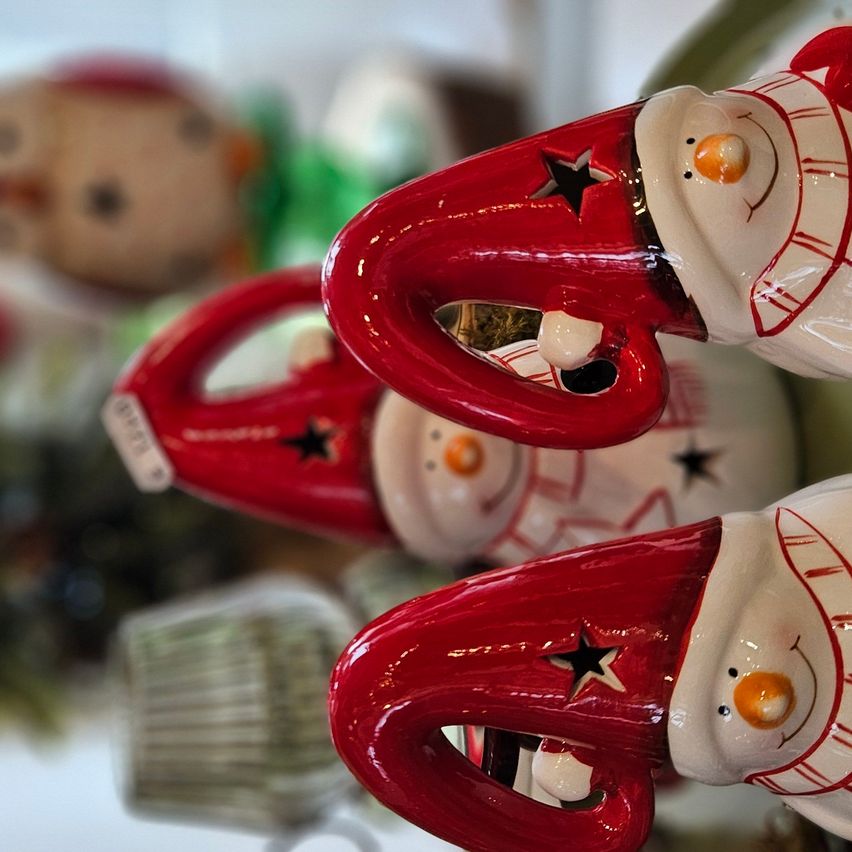 Drei Keramik-Schneemänner mit roten Mützen und weißen Schals stehen eng beieinander. Jeder hat einen schwarzen Stern auf seiner Mütze. Im Hintergrund sind verschwommene Weihnachtsdekorationen zu sehen.