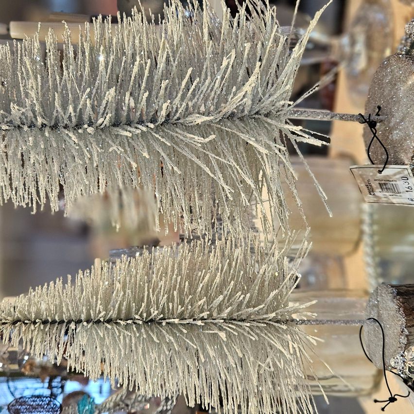 Eine Nahaufnahme eines dekorativen, silberstaubbedeckten Tannenzweigs mit glänzendem, metallische Finish, präsentiert vor einem verschwommenen Hintergrund von Weihnachtsschmuck.