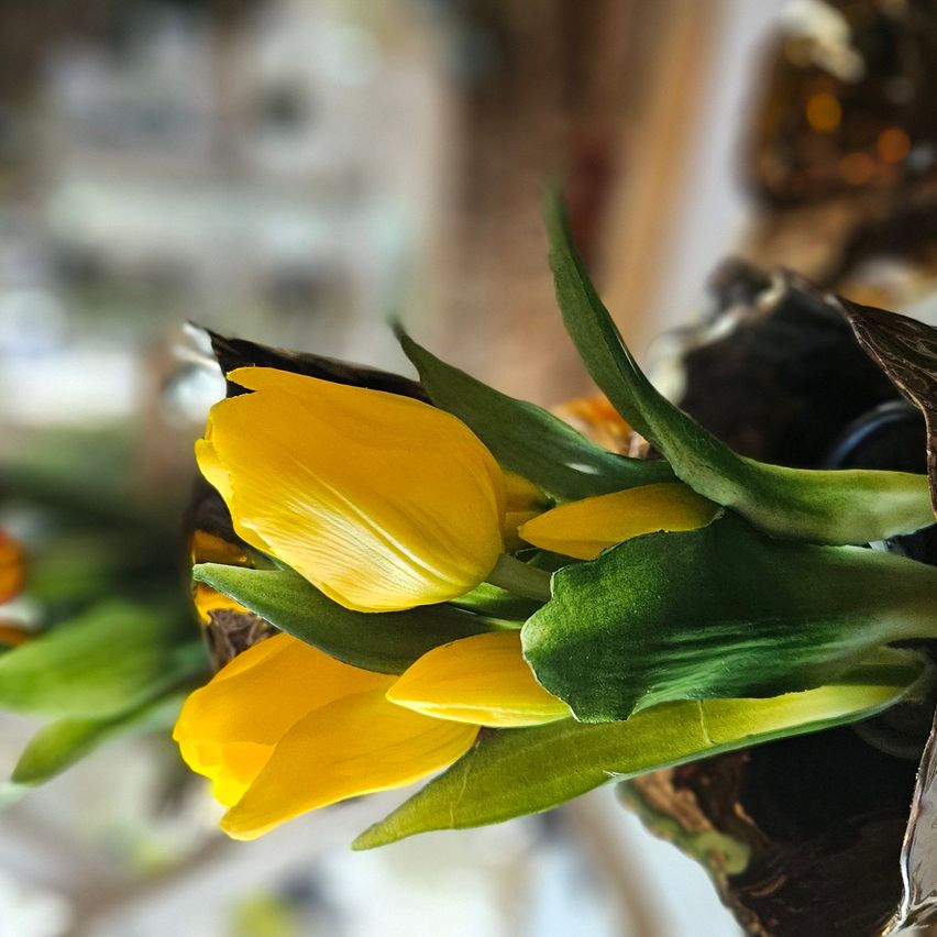 A close-up of a bouquet of yellow tulips in a vase, with green leaves and a blurry background.