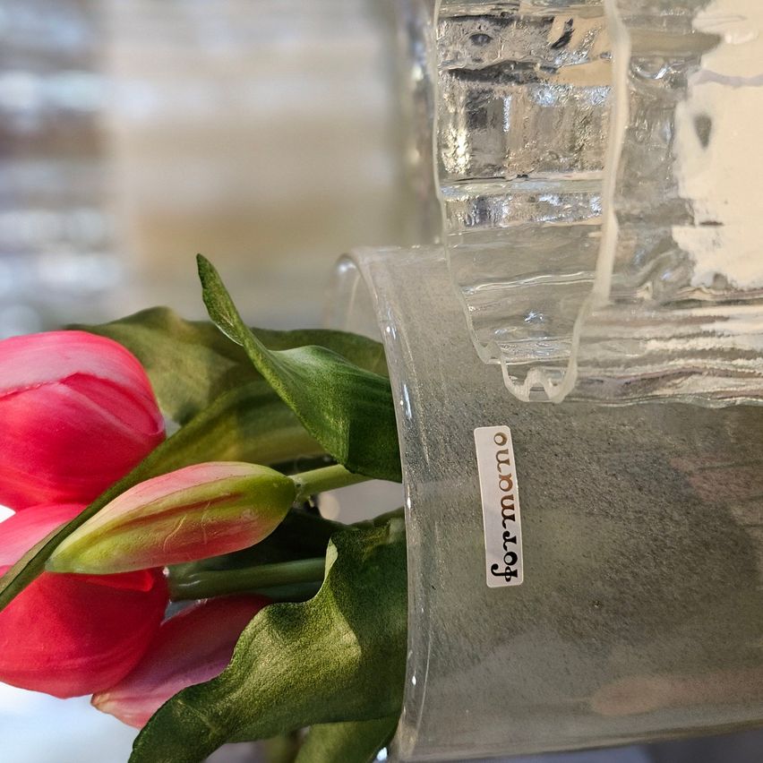 A close-up of pink tulips with green leaves in a clear glass vase with the word 'formmaro' on it.