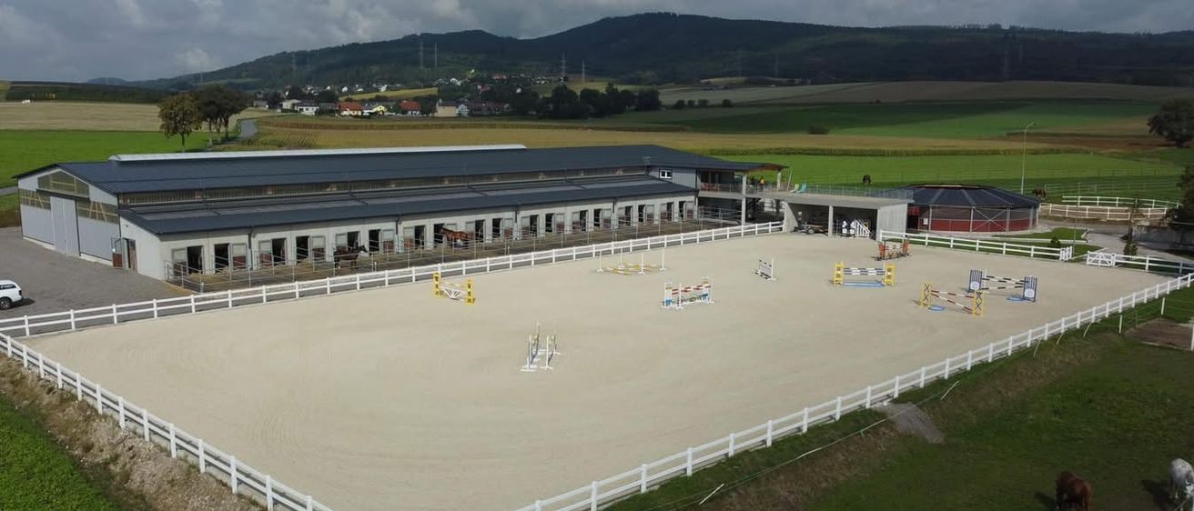 Luftaufnahme einer Reitanlage mit Sandarena, umgeben von weißen Zäunen. Gebäude mit Ställen und einer Veranda sind zu sehen. Ein grünes Feld und Berge bilden den Hintergrund.