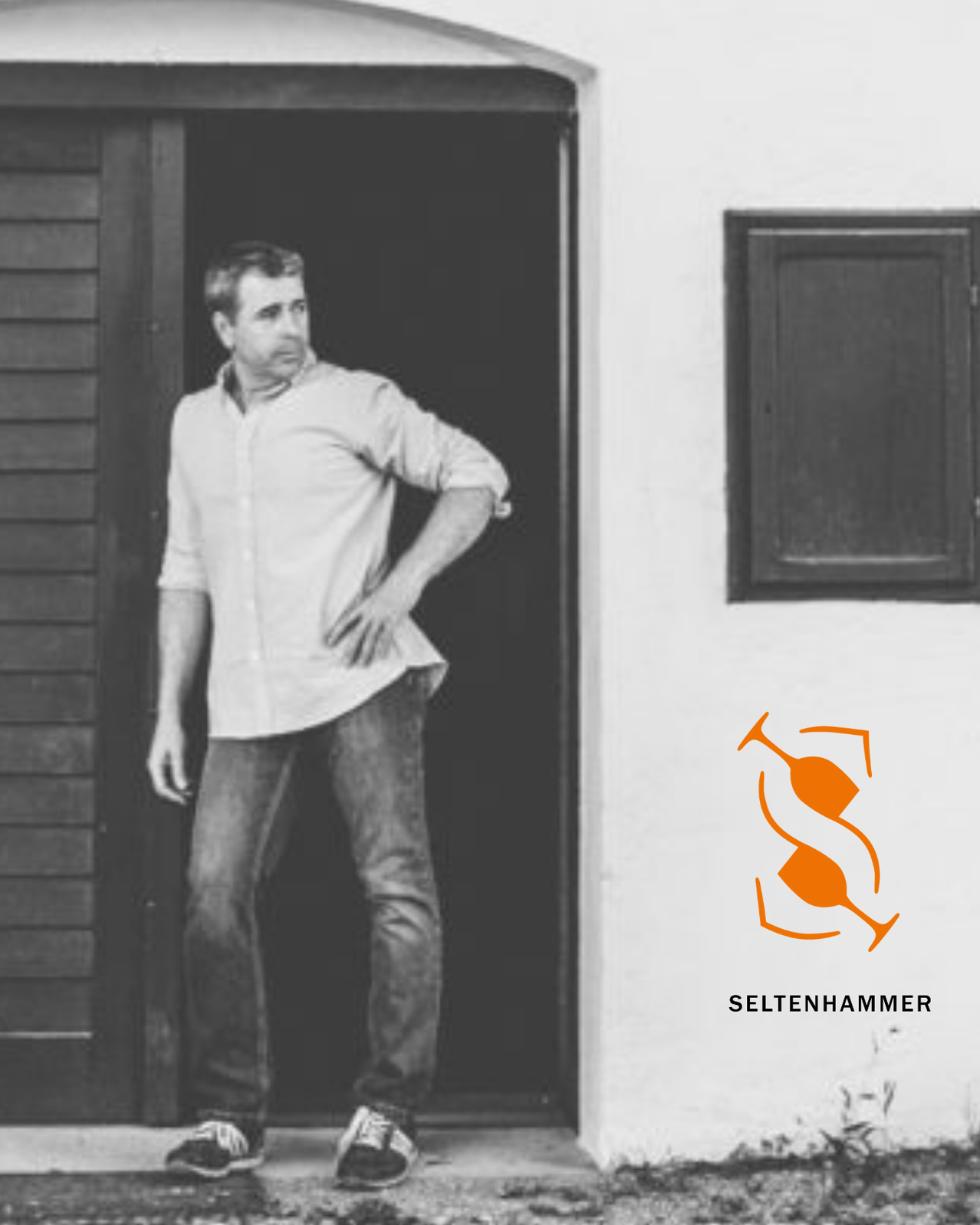 Ein Mann steht in der Tür eines Gebäudes, die Hände auf den Hüften. Die Tür ist aus Holz und das Hemd des Mannes ist weiß. In der Ecke befindet sich ein Logo.