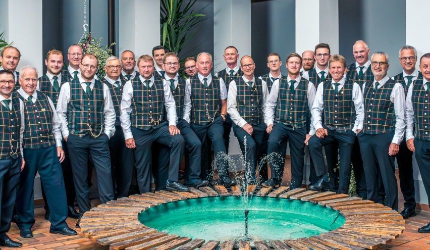 Eine Gruppe von Männern in karierten Westen und Krawatten posiert für ein Foto neben einem Brunnen.