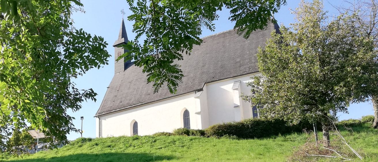 Eine kleine weiße Kirche mit grauem Dach und einem Kirchturm steht auf einem grünen Hügel unter einem klaren blauen Himmel.