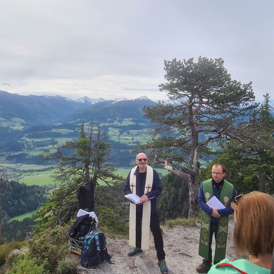 Zwei Männer in Gewändern stehen auf einem Berggipfel mit einer Frau und blicken auf ein Tal und schneebedeckte Berge. In der Nähe liegen ein Rucksack und ein Buch auf dem Boden.