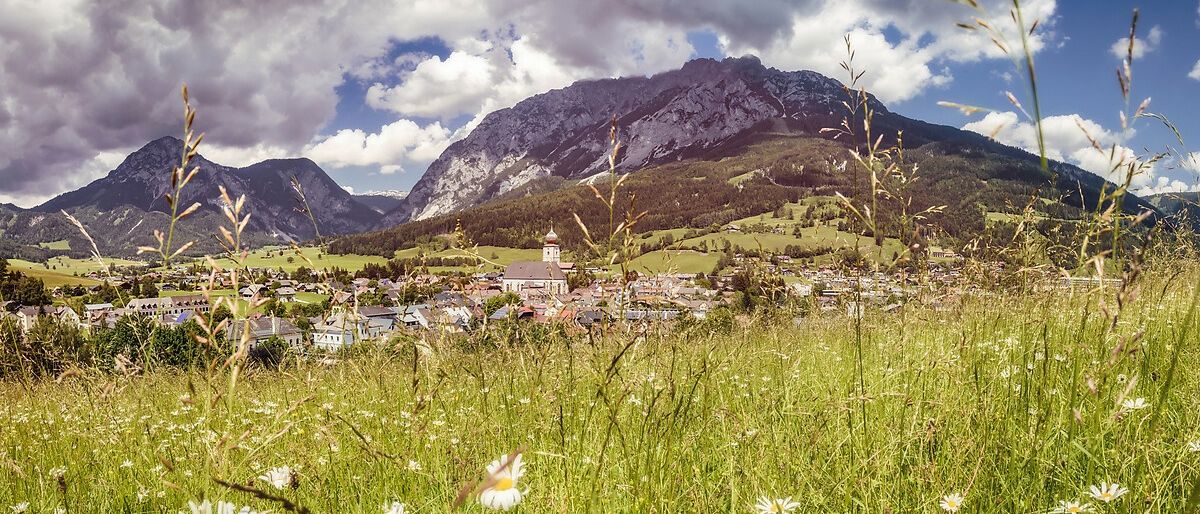 Ein malerisches Dorf in den Bergen mit einer Kirche in der Mitte, umgeben von üppigen grünen Feldern und Wildblumen unter einem bewölkten Himmel.