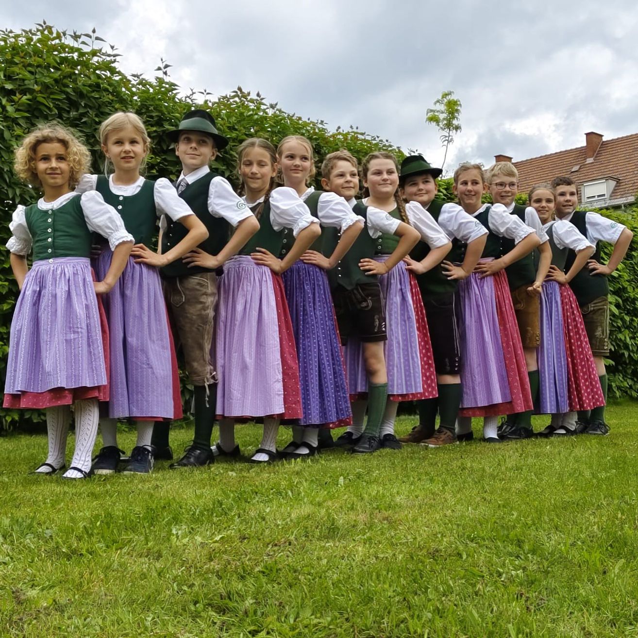 Eine Gruppe von Kindern in traditioneller Kleidung steht in einer Reihe auf einem Rasen, mit einem Haus und Büschen im Hintergrund.