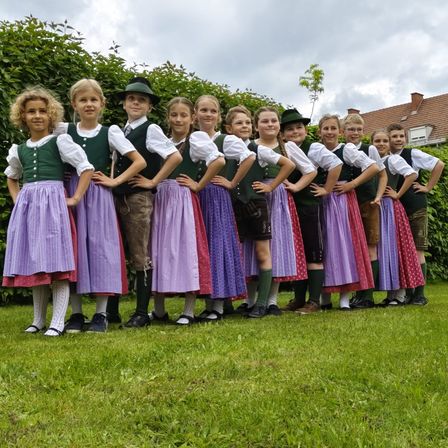 Eine Gruppe von Kindern in traditioneller Kleidung steht in einer Reihe auf einem Rasen, mit einem Haus und Büschen im Hintergrund.