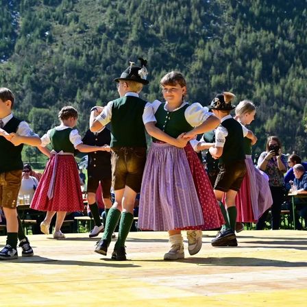 Eine Gruppe von Kindern in traditioneller Kleidung tanzt auf einer Bühne mit Bergen im Hintergrund. Einige Erwachsene schauen zu.