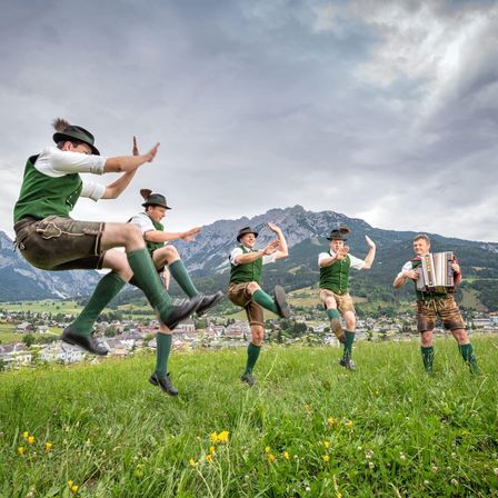 Fünf Männer führen einen traditionellen Tanz auf einem Grashügel auf, tragen grüne Westen, braune Hosen und schwarze Schuhe. Ein Mann spielt Akkordeon. Dahinter sind Berge und ein Dorf unter einem bewölkten Himmel sichtbar.