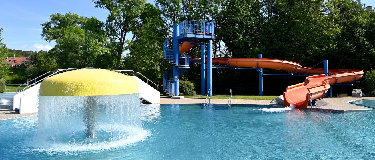 Außenpool mit blauer Rutsche und gelbem Wasserbrunnen, umgeben von üppiger Grünfläche. Bäume säumen den Hintergrund unter einem klaren blauen Himmel.