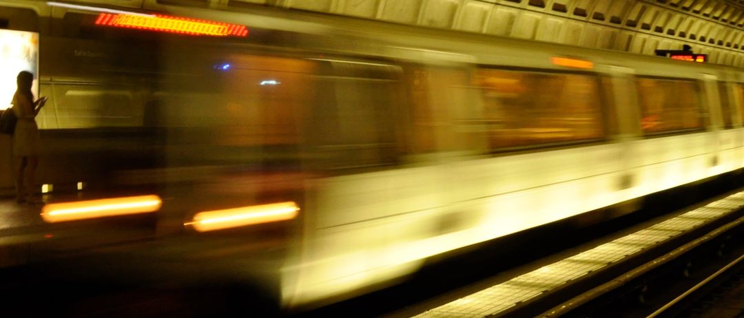 Ein U-Bahn-Zug bewegt sich mit verwischtem Effekt auf einer Schiene, während zwei Personen auf dem Bahnsteig stehen.