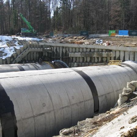 Eine Baustelle mit großen Betonrohren und einem Kran. Das Gebiet ist von einem Wald umgeben und es liegt Schnee auf dem Boden.