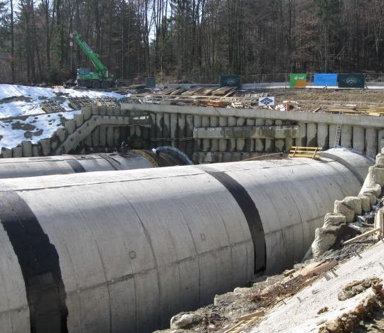 Eine Baustelle mit großen Betonrohren und einem Kran. Das Gebiet ist von einem Wald umgeben und es liegt Schnee auf dem Boden.