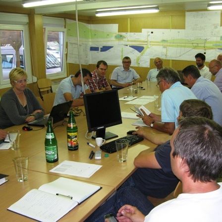 Eine Gruppe von Menschen sitzt in einem Konferenzraum an einem Tisch und führt eine Diskussion. Papiere und Laptops sind vorhanden, eine große Karte ist an der Wand zu sehen.