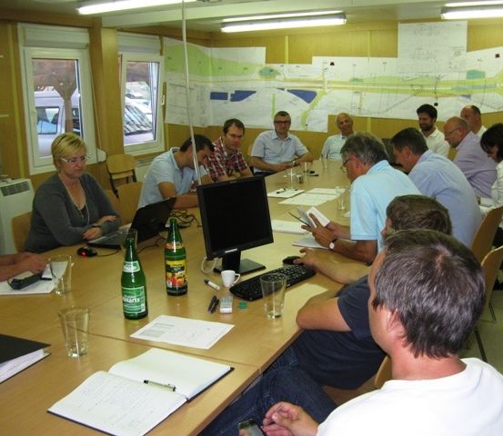 Eine Gruppe von Menschen sitzt in einem Konferenzraum an einem Tisch und führt eine Diskussion. Papiere und Laptops sind vorhanden, eine große Karte ist an der Wand zu sehen.