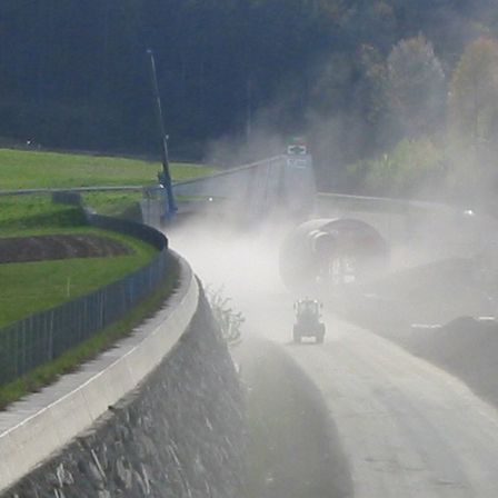 Ein Traktor fährt mit Nebel auf einer Straße, in der Nähe eines Grasfeldes und einer Betonbarriere. Bäume und eine Brücke sind im Hintergrund.