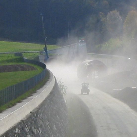 Ein Traktor fährt mit Nebel auf einer Straße, in der Nähe eines Grasfeldes und einer Betonbarriere. Bäume und eine Brücke sind im Hintergrund.