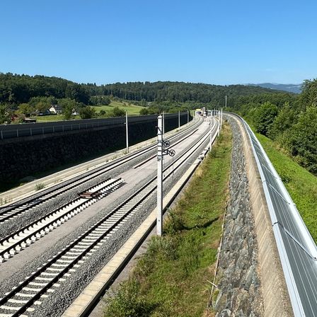 Eine erhöhte Eisenbahn mit zwei Gleisen wird von Steinmauern, grünem Gras und Solarpanels flankiert. Im Hintergrund befinden sich Bäume und Berge.