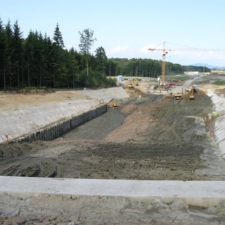 Eine Baustelle mit Erde, Beton und Maschinen. Bäume und ein Kran sind sichtbar.