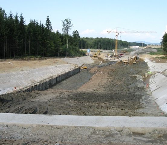 Eine Baustelle mit Erde, Beton und Maschinen. Bäume und ein Kran sind sichtbar.