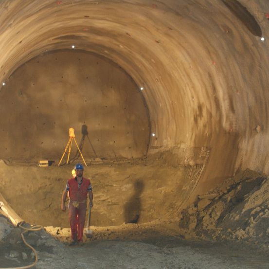 Ein Bauarbeiter in einem roten Overall steht in einem Tunnel, hält eine Schaufel und hat ein gelbes Stativ im Hintergrund. Die Tunnelwände sind mit Schmutz bedeckt.