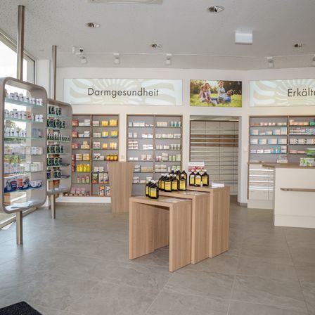 Ein Apothekeninnenraum mit mehreren Regalen, die mit verschiedenen Produkten gefüllt sind. Flaschen stehen auf dem Tresen. An den Wänden hängen Banner und Bilder von Menschen.