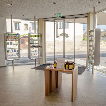 Ein leerer Raum mit einem Holztisch in der Mitte, auf dem mehrere Flaschen stehen. Zwei Glastüren und Regale mit verschiedenen Artikeln sind zu sehen. Draußen ist ein Gebäude und eine Straße durch die Glastüren zu erkennen.