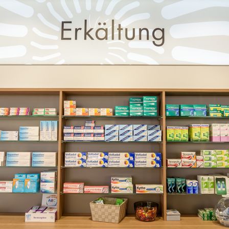 Eine Apotheke mit Regalen voller Medikamente und Produkten. Über dem Regal steht 'Erkältung'. Unten sind Boxen von Voltadol und Strepsils zu sehen. Ein Korb und eine Schüssel mit Süßigkeiten stehen auf dem Tresen.
