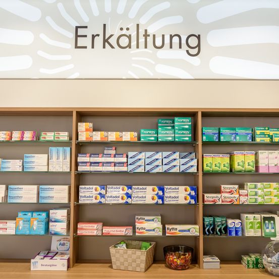 Eine Apotheke mit Regalen voller Medikamente und Produkten. Über dem Regal steht 'Erkältung'. Unten sind Boxen von Voltadol und Strepsils zu sehen. Ein Korb und eine Schüssel mit Süßigkeiten stehen auf dem Tresen.