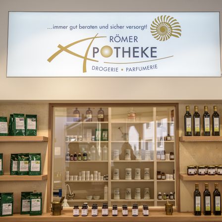 Eine Drogerie und Parfümerie mit einem Schild oben, das 'Römer Apotheke Drogerie Parfumerie' besagt. Flaschen und Gläser sind auf den Regalen ausgestellt.