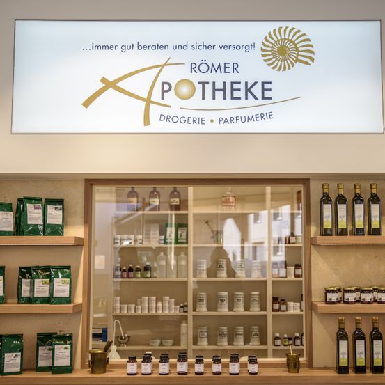 Eine Drogerie und Parfümerie mit einem Schild oben, das 'Römer Apotheke Drogerie Parfumerie' besagt. Flaschen und Gläser sind auf den Regalen ausgestellt.