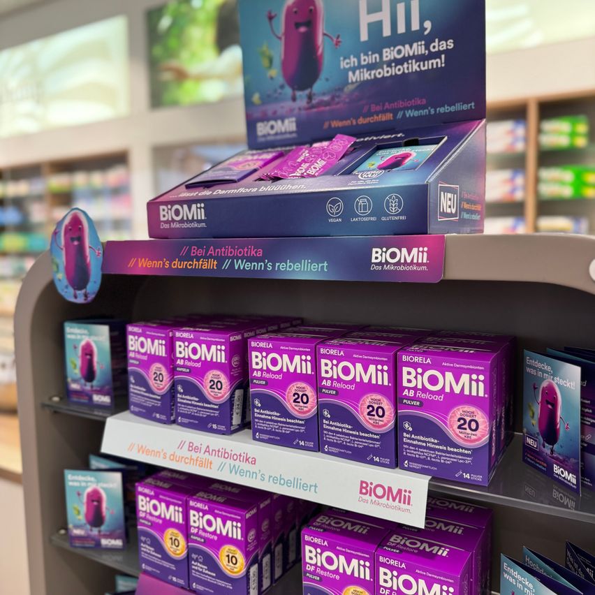 Ein Regal in einer Apotheke zeigt lilafarbene Schachteln von BIOMiI-Probiotika, mit einem Cartoon-Charakter auf der Vorderseite. Die Schachteln sind in Reihen angeordnet, wobei die oberste Reihe eine größere Display-Box zeigt.