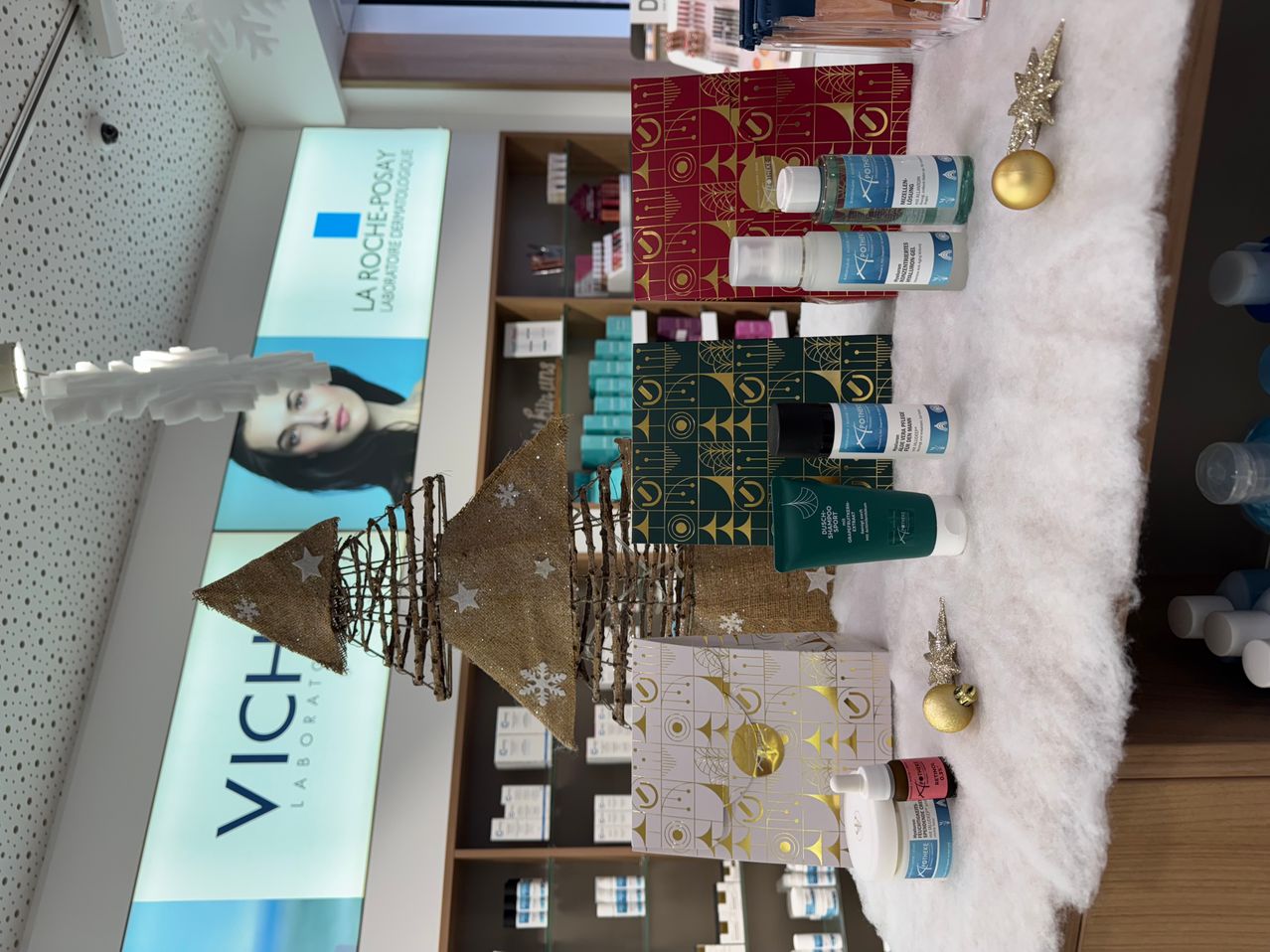 Ein Schönheitssalon-Display mit einem Weihnachtsbaum, verpackten Geschenken und Hautpflegeprodukten. Ein Banner mit der Aufschrift 'Vichy Laboratories'. Verschiedene Hautpflegeprodukte und Ornamente sind auf einer weißen Pelzoberfläche angeordnet.