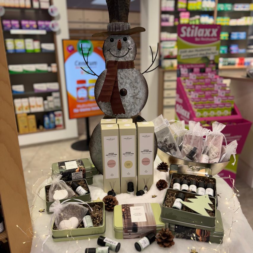 Ein Tischdisplay in einem Geschäft mit einem Schneemann und verschiedenen Geschenksets, einschließlich Kerzen, kleinen Flaschen und Tannenzapfen. Im Hintergrund sind Regale mit Produkten zu sehen.