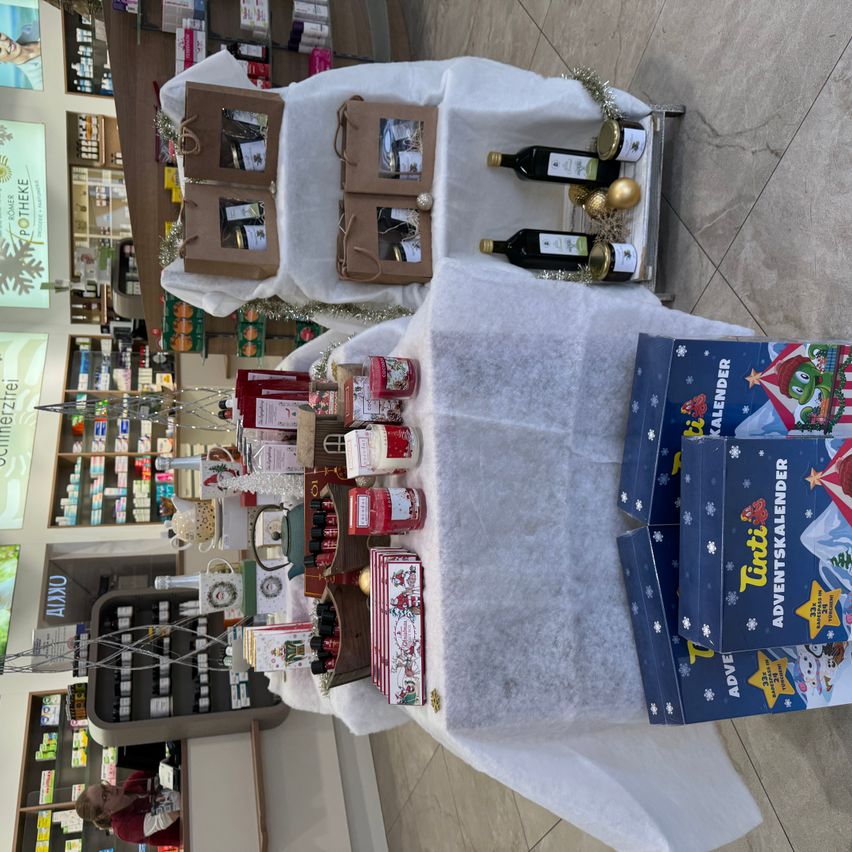 Eine Übersicht eines Ladens mit einem Tisch, der mit einem weißen Tuch bedeckt ist, mit verschiedenen Artikeln, darunter Geschenkboxen, Flaschen und Schachteln mit der Aufschrift 'Adventskalender'. Regale mit Produkten und einer Person im Hintergrund.