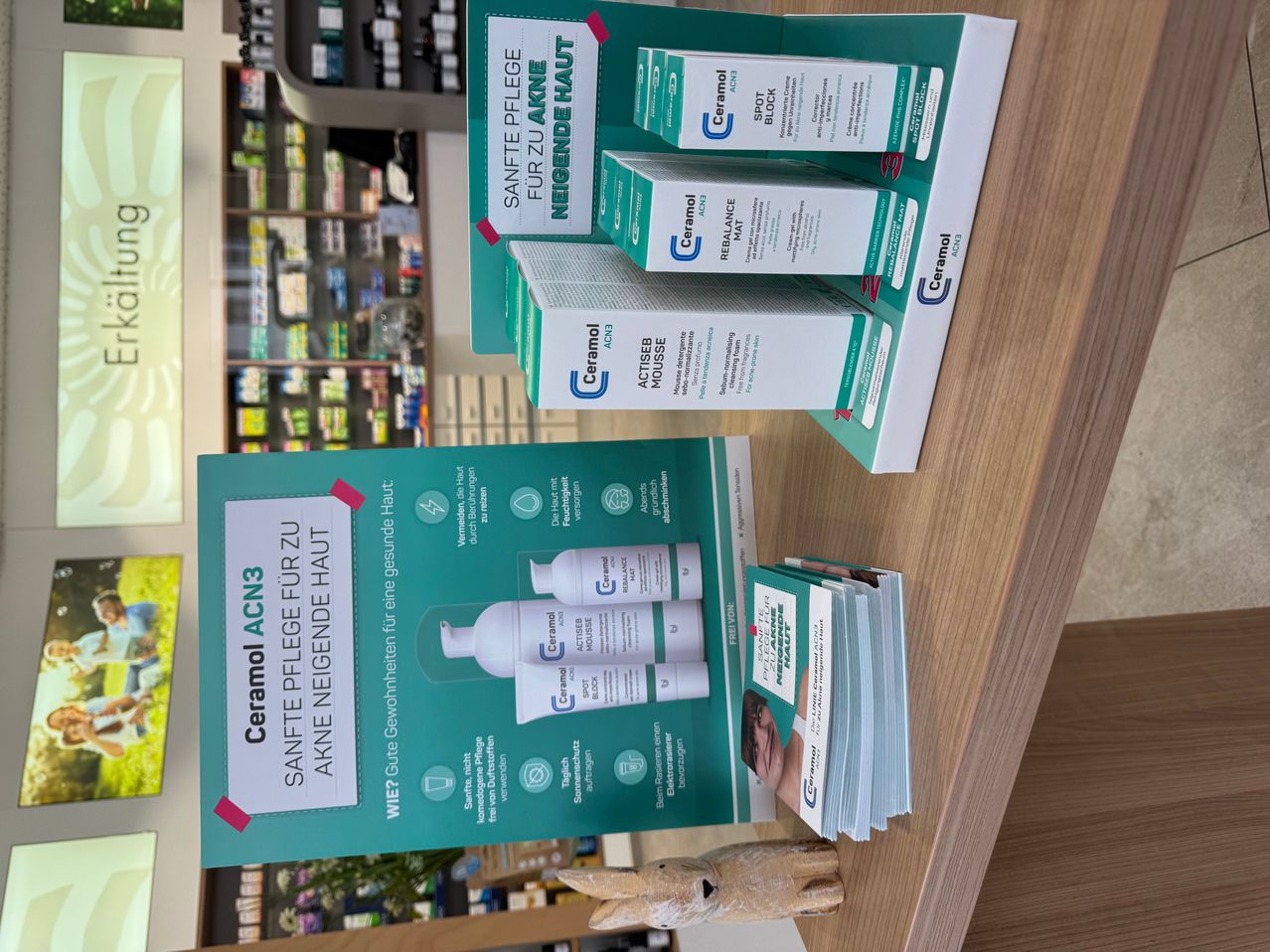 Mehrere Ceramol-Produkte, einschließlich Kartons und Broschüren, sind auf einer Holzoberfläche in einer Apotheke ausgestellt. Die Produkte sind gestapelt und zur Ausstellung positioniert.