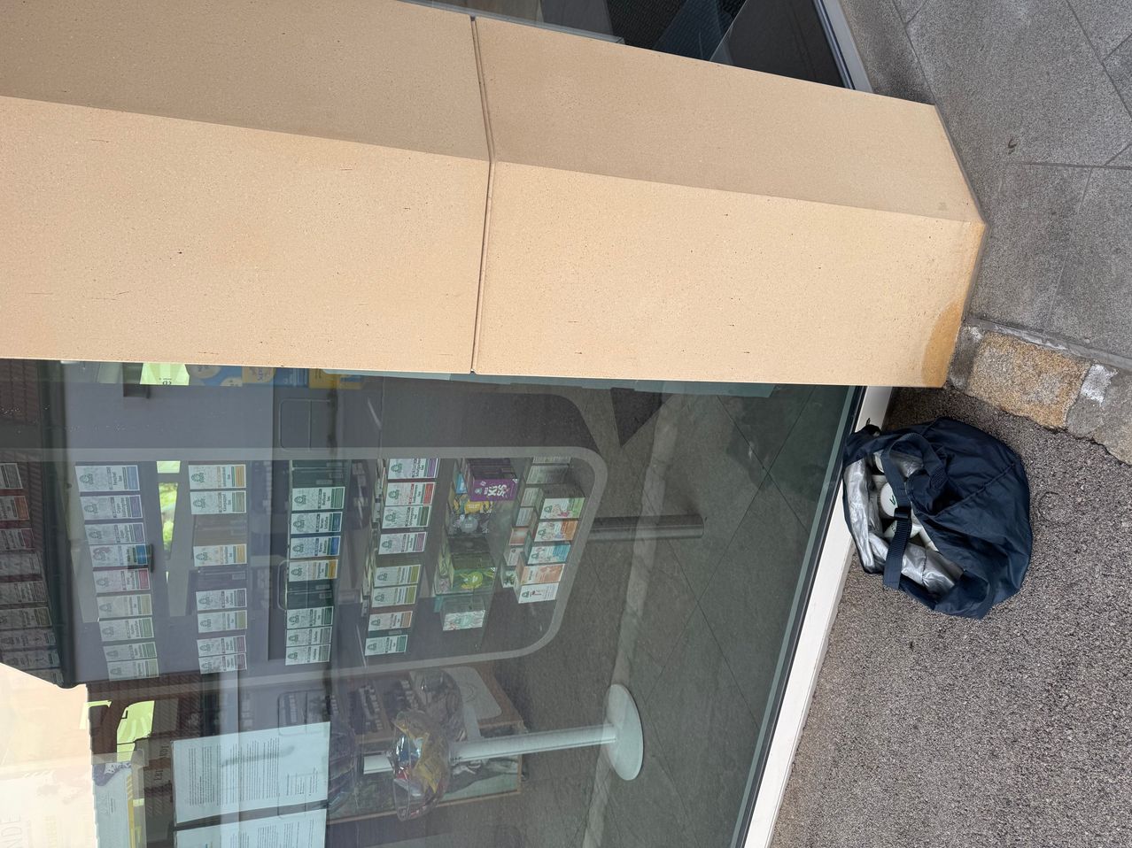 Atop a glass door, a cardboard box is positioned. Below it, a shelf with various items is visible through the glass. A blue bag rests beside the door.