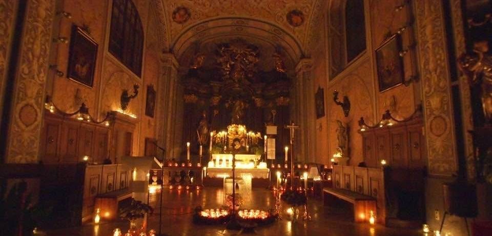 Ein kerzenbeleuchteter Altar in einer Kapelle mit kunstvollen Wanddekorationen, einem zentralen Kerzenständer und Bänken auf beiden Seiten. Die Wände sind mit gerahmten Bildern geschmückt.