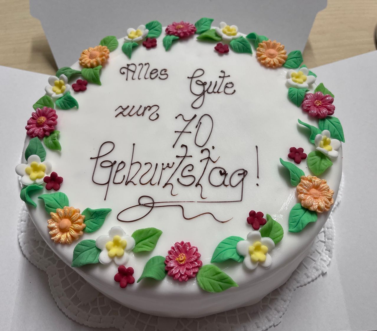 Ein weißer Kuchen mit bunten Blumen und Blättern am Rand, mit der Aufschrift 'Alles Gute zum 70. Geburtstag!' in Braun auf dem Kuchen.