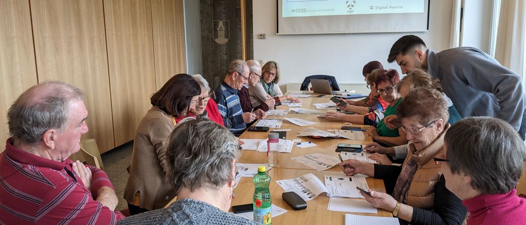 Eine Gruppe älterer Menschen sitzt um einen Tisch herum und ist mit einer Aktivität beschäftigt. Ein Beamer hinter ihnen zeigt eine Präsentation mit dem Titel 'Vorübung 1'. Jede Person hat Stift und Papier. Einige benutzen ihre Telefone. Flaschen und andere Gegenstände stehen auf dem Tisch.