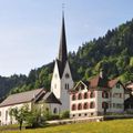 Eine Dorfkirche mit einem hohen Turm und einer Uhr liegt in einem grünen Tal, mit einem Haus in der Nähe und einem Berg im Hintergrund.