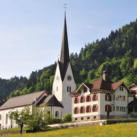 Eine Dorfkirche mit einem hohen Turm und einer Uhr liegt in einem grünen Tal, mit einem Haus in der Nähe und einem Berg im Hintergrund.