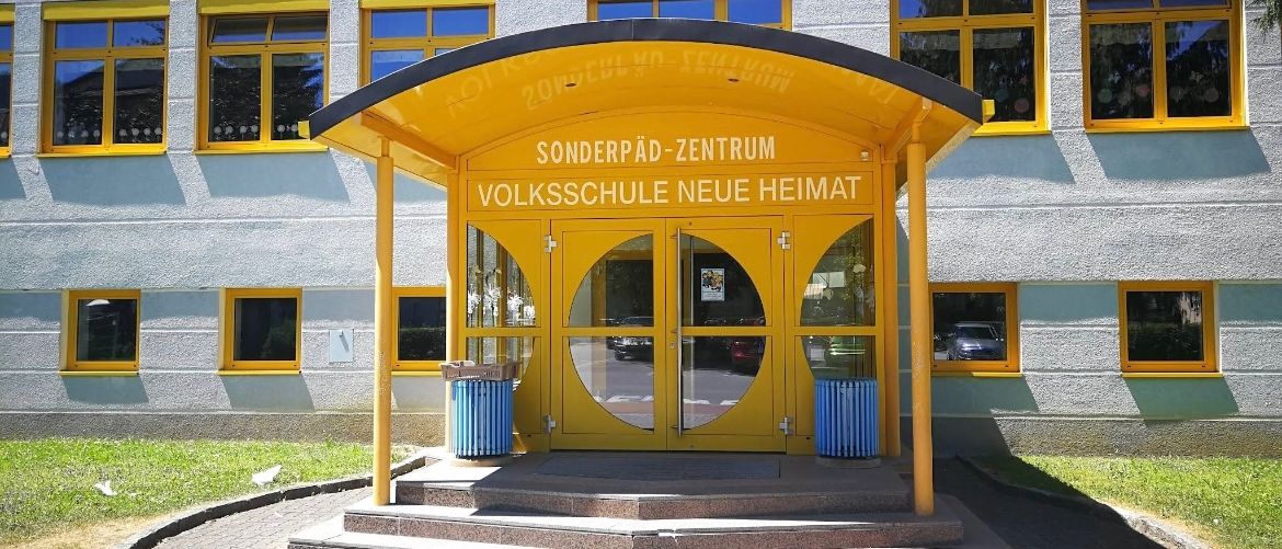 Der Eingang eines Gebäudes mit gelben Fenstern und einem gelben Dach hat zwei Türen. Das Schild steht Sonderpad-Zentrum Volksschule Neue Heimat.