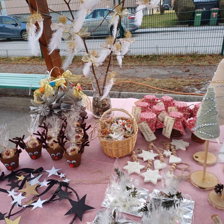 Ein mit einem rosa Tuch bedeckter Tisch hat verschiedene Gegenstände wie Gläser, einen Korb und festliche Dekorationen. Es gibt renntierartige Gegenstände und Sterne auf dem Tisch. Ein Baumzweig mit Federn und ein Vasen steht links. Hinter einem Zaun gibt es eine Bank und geparkte Autos.