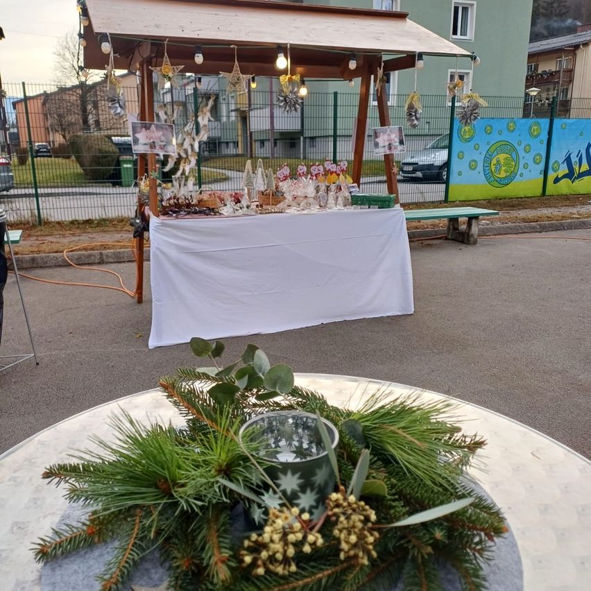 Ein Außenmarktstand ist mit einem Vordach aufgestellt, geschmückt mit Lichtern und Feiertagsdekoration. Ein Tisch, bedeckt mit einem weißen Tuch, zeigt verschiedene Süßigkeiten an. In der Nähe gibt es eine Bank und ein farbenfrohes Banner. Die Szene ist in einem gepflasterten Bereich, umgeben von einem grünen Zaun.